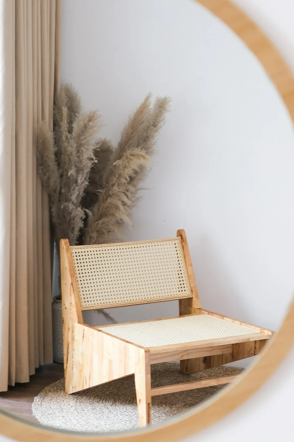 Round rattan mirror reflecting a rattan chair and pampas grass